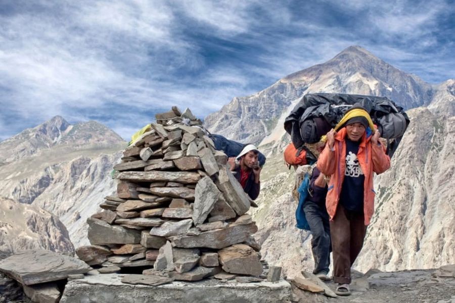 Trekkers with heavy packs crossing high mountain pass with stone cairn and snow-capped peaks